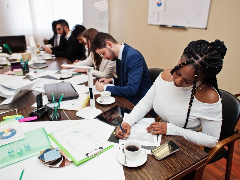 Multiracial business team addressing meeting around boardroom table, working together and write something on papers.